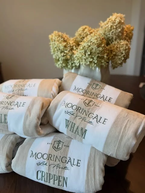 Stack of folded Mooringale Lake Martin beach towels with an anchor crest and embroidered family names