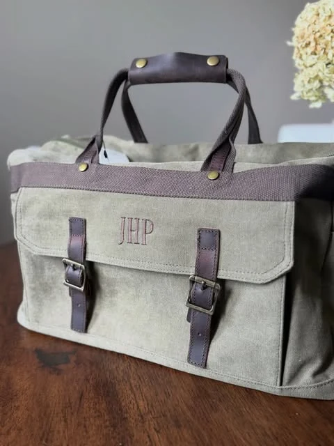 An olive waxed canvas weekender duffel bag with brown leather straps, brass hardware, and a burgundy monogram reading JHP